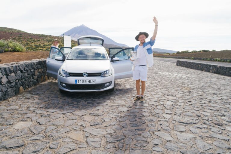 Cheerful male in white clothes standing on cobblestone near car and greeting friend while resting with wife in countryside